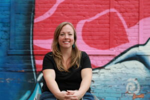 Circular portrait of Dessa sitting in her wheelchair in front of a mural with different color pinks & blues in pattern. She smiles in a black tee shirt with blue dangling earrings. She has long blonde hair and light skin. Her arms come together in front of her lap with her fingers interlaced.