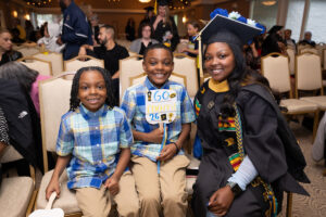 A graduate in a black cap and gown decorated with blue flowers smiles alongside her two young sons, who are seated in chairs at a ceremony. The older boy holds a handmade sign reading "Go Mom! 26."