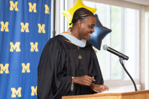 A graduate in a black cap and gown with a light blue doctoral hood smiles while speaking at a podium microphone. A University of Michigan step-and-repeat backdrop and gold and navy star-shaped balloons are visible behind them.