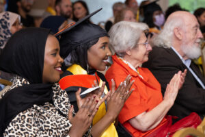 A graduate wearing a black cap and gown with a yellow and orange doctoral hood claps and smiles while seated at a ceremony. Beside her is a woman in a black hijab and leopard-print top, and two older adults in the seats next to them also applaud.