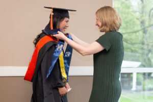 A staff member in a green dress smiles as she places a blue and gold CEW+ stole around the neck of a beaming graduate in a black cap and gown with an orange stole. They face each other near a window with trees visible outside.
