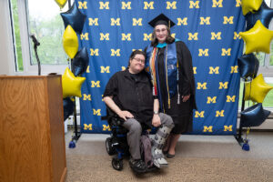 A graduate in a black cap and gown with a blue CEW+ stole and colorful honor cords poses with a supporter seated in a power wheelchair, both smiling in front of a University of Michigan step-and-repeat backdrop decorated with navy and gold star-shaped balloons.