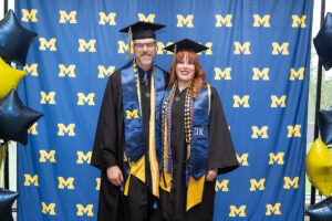 Two graduates in black caps and gowns with blue and gold University of Michigan stoles pose together in front of a U-M step-and-repeat backdrop flanked by navy and gold star balloons.