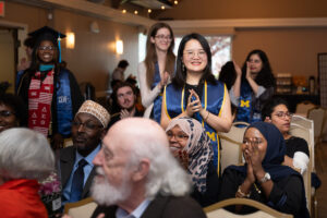 Graduates and guests applaud during a ceremony in a warmly lit banquet room. In the foreground, a smiling graduate wearing a blue University of Michigan CEW+ stole claps enthusiastically while standing among seated attendees.