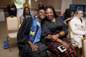 A graduate in a black cap and gown with a blue CEW+ stole leans in close to a smiling supporter seated beside them at a ceremony, both laughing and looking at the camera.
