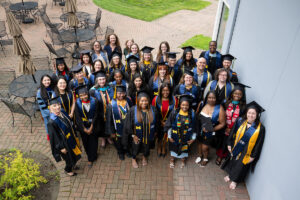 An aerial view of a large group of University of Michigan graduates in black caps and gowns with blue and gold CEW+ stoles posing together on a brick outdoor patio, looking up toward the camera with smiles.