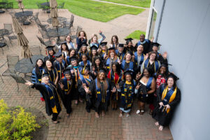 An aerial view of a large group of University of Michigan graduates in black caps and gowns with blue and gold CEW+ stoles, laughing, waving, and making peace signs on a brick outdoor patio. Patio furniture and green grass are visible in the background.