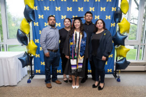 A graduate in a black cap and gown with a blue CEW+ stole and a colorful sash stands in the center of a group of four family members, all smiling in front of a University of Michigan step-and-repeat backdrop with navy and gold star balloons.
