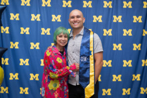 A graduate wearing a blue and gold CEW+ stole puts his arm around an older woman with short green hair in a bright floral top. Both smile at the camera in front of a University of Michigan step-and-repeat backdrop.