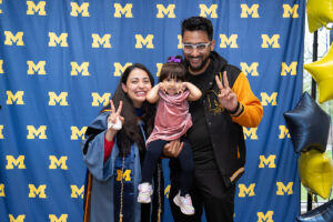 A graduate in a blue doctoral gown and gold honor cords flashes a peace sign alongside a man holding a toddler in a pink outfit, all three smiling in front of a University of Michigan step-and-repeat backdrop with gold and navy star balloons.
