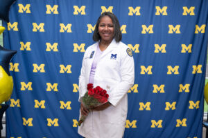 A graduate wearing a white University of Michigan–Flint lab coat holds a bouquet of red roses and smiles in front of a University of Michigan step-and-repeat backdrop with navy and gold balloons visible at the edges.